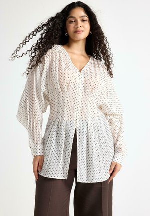Langærmet, hvid bluse med sorte polkaprikker, plisseret front, v-udskæring og knaplukning, kombineret med brune bukser.