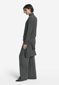 Grauer gestrickter Pullover mit hohem Kragen, der Kabelmuster aufweist. Kombiniert mit weiten grauen Hosen, die beide aus einem weichen, strukturierten Stoff gefertigt sind.