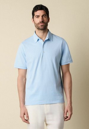 Man die een lichtblauw poloshirt met korte mouwen en een witte broek draagt, staand tegen een beige achtergrond, naar voren kijkend met een neutrale gezichtsuitdrukking.