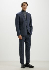 Completo a quadri blu navy con vestibilità su misura, abbinato a un dolcevita grigio. Completato con eleganti scarpe nere e dettagli in texture discreti.