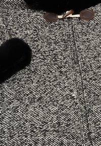 Svart och vit fiskbensmönstrad tyg med en mjuk textur, med svarta faux fur-accenter och bruna läderknäppningar.