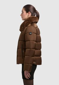Brun pufferjakke med en faux fur-krave, quiltet design, sidelommer med lynlås og logo-èmblem på skulderen. Tekstureret livsgangsstyle.