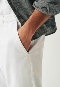 Pantalons en lin blancs avec une poche latérale, assortis à une chemise noire et blanche texturée. Main visible reposant dans la poche.