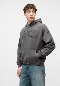 Νεαρός άνδρας που φοράει ξεθωριασμένη μαύρη μπλούζα με κουκούλα και το κείμενο "YAH", με το ένα χέρι στην τσέπη, συνδυασμένη με ανοιχτόχρωμα τζιν με σχέδιο patchwork, κοιτάζοντας προς τα πλάγια.