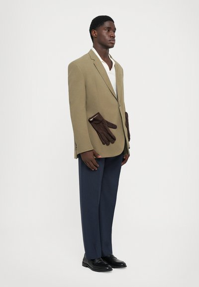 Beiger Blazer mit braunen Lederhandschuhen in den Taschen, weißem Hemd darunter, marineblauen Stoffhosen und schwarzen Schuhen. Schlankes, strukturiertes Design, texturierte Materialien.