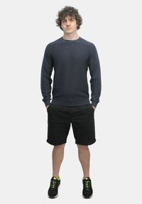 Maglione blu navy a maglia, con colletto rotondo, indossato con pantaloni corti neri. Modello in sneakers nere con dettagli al neon, in piedi con le mani nelle tasche.