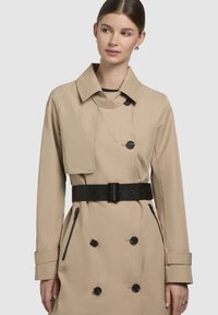 Beige trench coat featuring a double-breasted design, black buttons, and a wide black belt. Includes a folded collar and side zip pockets.