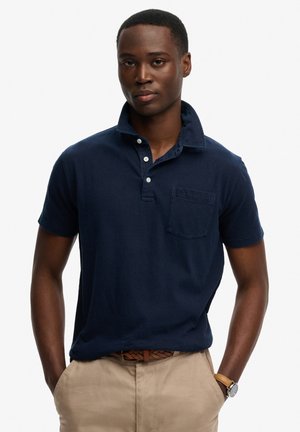 Joven con una camisa polo azul marino con un bolsillo en el pecho, pantalones beige, un cinturón trenzado marrón y un reloj de pulsera, con las manos en los bolsillos.