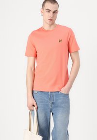 Korallfärgad bomullströja med rund hals och korta ärmar, med en liten gul logotyp på bröstet. Matchas med blå denimjeans.