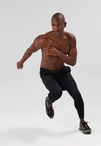 Sportininkas juodais šortais ir juodomis leggingomis bėga. Jis turi raumeningą kūną, trumpus plaukus ir nešioja juodus bėgimo batus su žaliais akcentais.