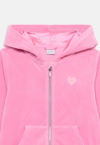 Sudadera con cremallera de terciopelo rosa que presenta un acento de purpurina en forma de corazón, dos bolsillos frontales y una capucha forrada con una textura suave.