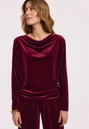 Kobieta z falującymi, jasnobrązowymi włosami, ubrana w długosłowy top z głębokiego burgundowego aksamitu i pasujące spodnie, patrząca w lewo na jednolitym tle.