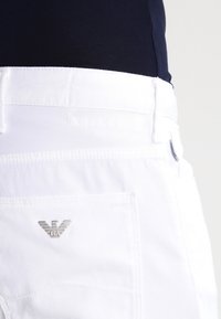 White pants with a smooth texture, featuring a back pocket with a silver logo accent and a contrasting colored waistband.