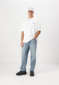 White oversized T-shirt with logo, light blue jeans, and black sturdy sneakers. Accessories include a light beige cap. Simple, casual outfit.