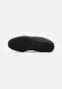 Schwarze Sneaker-Sohle mit einem strukturierten Muster, das kreisförmige Griffe und eine glatte Kante aufweist. Das Material scheint Gummi mit einem leichten Glanz zu sein.