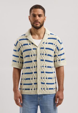 Man wearing a short-sleeve knit cardigan with cream and blue stripes over a white shirt, paired with light blue jeans.