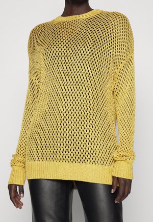 Person trägt einen lockeren, gelben, offen gestrickten Pullover über schwarzen Lederhosen, dargestellt von den Schultern bis zur Hüfte vor einem schlichten Hintergrund.