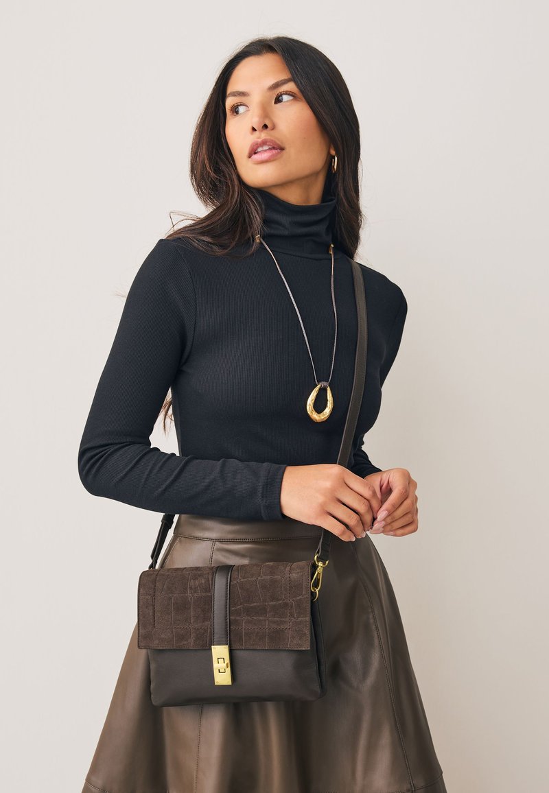 Femme aux longs cheveux portant un col roulé noir, une jupe en cuir marron, un collier pendentif en or, et portant un sac bandoulière marron avec une fermeture dorée.