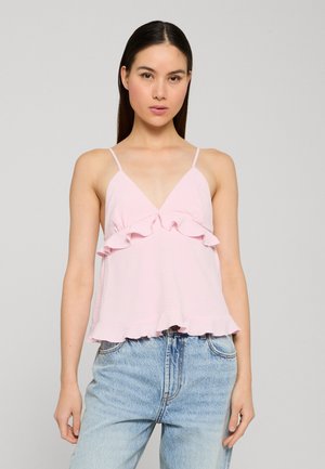 Femme aux cheveux raides et foncés portant un haut sans manches rose clair à volants et un jean taille haute bleu clair sur un fond uni.