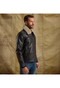 Hnedá kožená bunda s ovčím golierom, zipsom na uzavretie a bočnými vreckami. Má priliehavý pás a hladkú textúru.