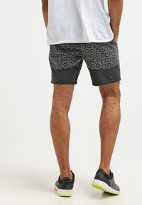 Man standing wearing black patterned shorts, white t-shirt, and black and gray sneakers with lime green soles, on plain light background.