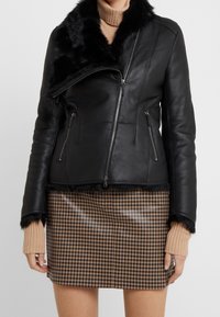 Černá kožená bunda s límcem z umělé kožešiny, zapínáním na zip a kapsami, kontrastující s sukní v houndstooth vzoru a ribovanými béžovými rukávy.