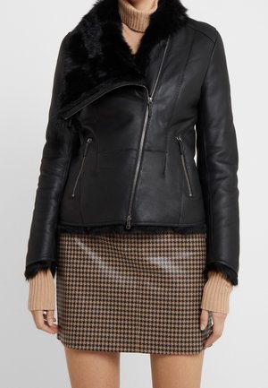 Czarna skórzana kurtka z futrzanym kołnierzem, zapinana na zamek, z kieszeniami, w zestawieniu z spódnicą w wzór houndstooth oraz beżowymi, prążkowanymi rękawami.