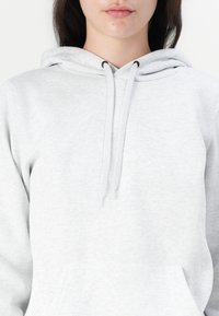 Femme portant un sweat à capuche gris clair avec poche avant et cordons, montrée des épaules aux lèvres sur fond blanc.