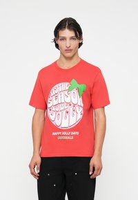Tricou din bumbac roșu cu un grafic alb rotund care spune „Sezonul să fim veseli”, accentuat de o fundă verde pe partea stângă.