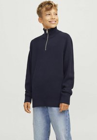 Mornarška zip-up jopica z visokim ovratnikom in rebrasto teksturo. Dolgi rokavi z manšetami. V kombinaciji s svetlo modrimi jeans hlačami.