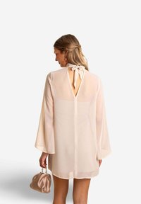 Robe beige clair en tissu transparent avec de longues manches évasées, un détail en forme de clé au dos et un lien en ruban au cou, accompagnée d'un sac doux.