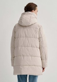 Giubbotto imbottito beige chiaro con cappuccio, caratterizzato da una texture trapuntata e cuciture verticali; lunghezza vita con tasche laterali e vestibilità comoda.