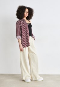 Camicia a righe bordeaux e bianca con bottoni sopra una canotta nera, abbinata a pantaloni a gamba larga crema e sneakers platform bianche.