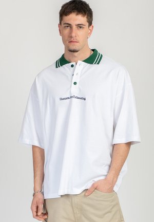 Giovane uomo con capelli corti indossa una polo bianca oversize con colletto verde e scritta "Humans Are Exhausting", pantaloni beige e braccialetto d’argento.