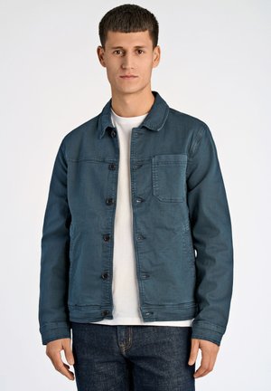 Dunkelblaue Jeansjacke mit knöpfbarem Verschluss, spitzem Kragen und einer Brusttasche. Über einem weißen T-Shirt getragen, kombiniert mit dunklen Jeans.