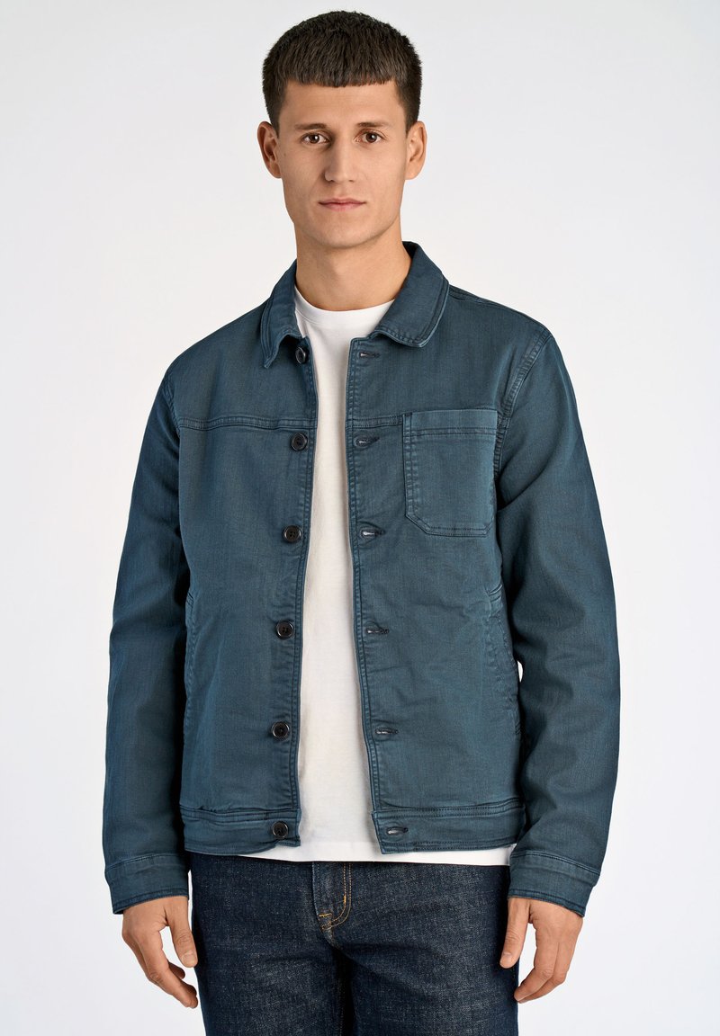 Tummansininen farkkutakki, jossa on napitettava etuosa, terävä kaulus ja yksi rintatasku. Päälle puettuna valkoinen t-paita, yhdistettynä tummiin farkkuihin.