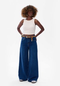 Biały prążkowany crop top, niebieskie szerokie dżinsy, brązowy pasek z srebrnymi elementami i szare buty. Modelka stoi z rękami na biodrach.