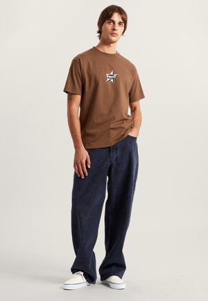Brown cotton t-shirt with a black and white star logo. Paired with loose-fitting dark blue denim jeans and white sneakers.