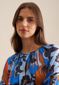Langärmlige Bluse mit blauem Grund, die große Blumenmuster in Orange und dunklen Farben aufweist; weicher Stoff mit gerafftem Ausschnitt.