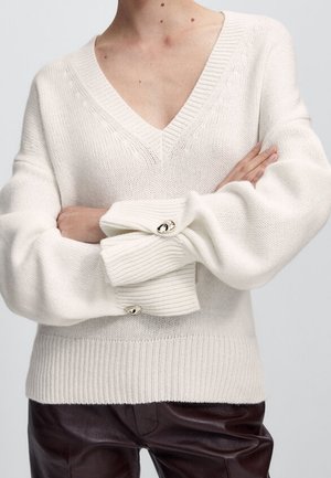 Personne portant un pull en maille crème oversize avec un profond col en V et des poignets boutonnés, associé à un pantalon en cuir marron foncé.