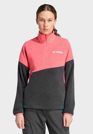 Vrouw met bruin haar in een paardenstaart draagt een roze en donkergrijze Terrex fleecejas met een kwart ritssluiting en donkere broek, staand tegen een effen achtergrond.