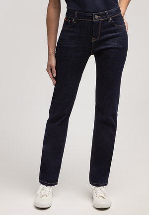 Kobieta w ciemnoniebieskich prostych dżinsach i białych trampkach, stojąca z jedną ręką na boku, a drugą na biodrze.