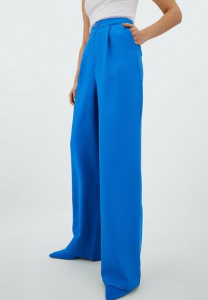 Femme portant un pantalon large taille haute bleu vif, assorti à des chaussures pointues de la même couleur, et un haut blanc sans manches.
