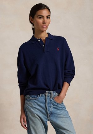 Sötét hajú nő alacsony lófarokkal, sötétkék hosszú ujjú pólóingben piros logóval és világoskék farmerral, egyszínű háttér előtt áll.