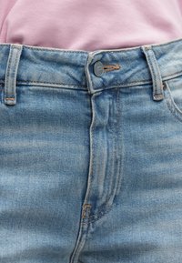 Τζιν παντελόνι από ανοιχτό μπλε τζιν ύφασμα σε ίσια γραμμή, με κούμπωμα κουμπιών μπροστά, ραφές σε χάλκινο χρώμα και ελαφρώς ξεθωριασμένη υφή.