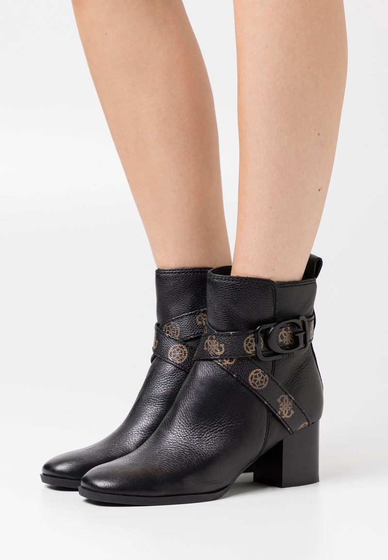 Bottes noires en cuir à la cheville avec une finition texturée, présentant une sangle décorative avec un motif logo et un talon carré.