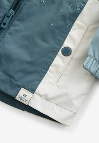 Gros plan sur le coin d'une veste bleu canard et beige avec fermeture éclair, bouton-pression étiqueté "NEXT ESTABLISHED 1982" et étiquette en tissu indiquant "OKAY" avec une étoile.