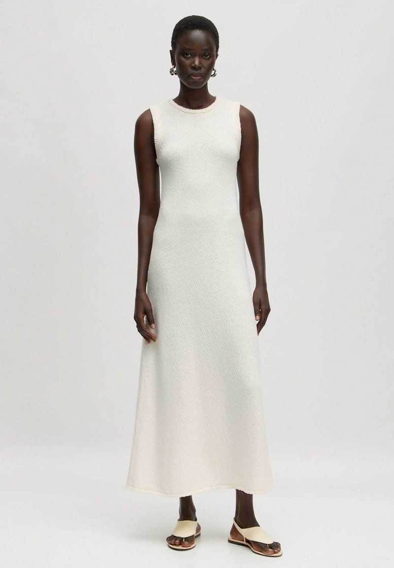 Gebreide witte mouwloze maxi jurk met een textuurafwerking, ronde halslijn en een rechte silhoët, gecombineerd met beige platte sandalen.