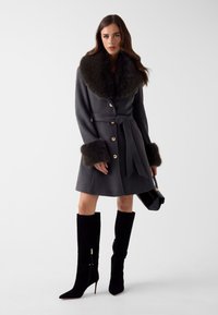 Grijze wollen jas met een grote bontkraag en nep bontmanchetten, met een ceintuur in de taille, gecombineerd met zwarte laarzen tot boven de knie en een kleine zwarte handtas.