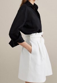 Donna che indossa una blusa nera a maniche lunghe infilata in una gonna bianca a vita alta con una cintura larga e pieghe, mano in tasca.